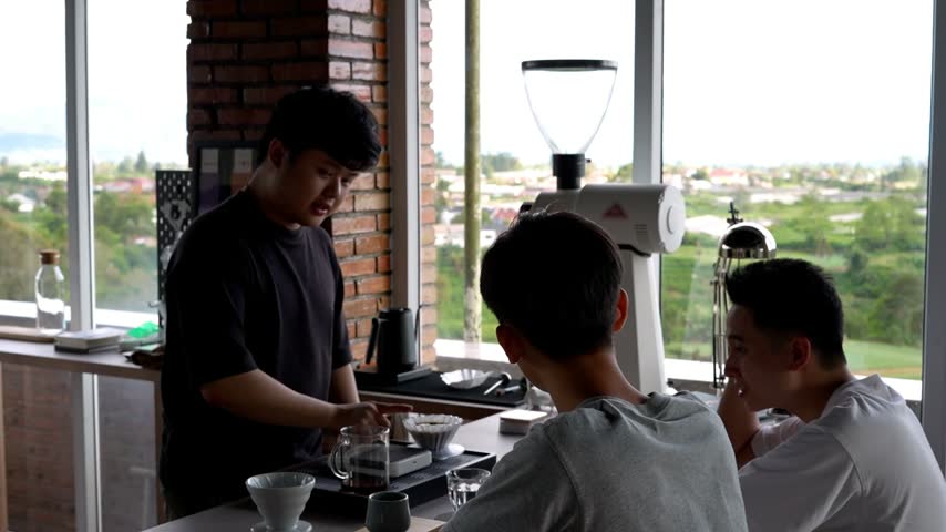 A friendly Asian barista explaining the manual coffee brewing process to two young male customers sitting at a cafe counter with a scenic window view