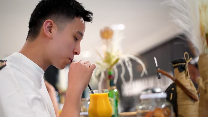 Side profile of a young Asian man casually sipping a bright yellow juice drink through a straw at a cafe or bar counter