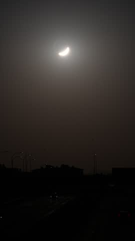 Partial solar eclipse creating a crescent sun in the hazy sky above a dark city silhouette