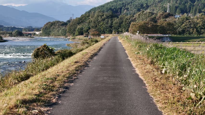 A paved path stretches past grasses and riverside trees with a flowing river on one side and wooded hills beyond.