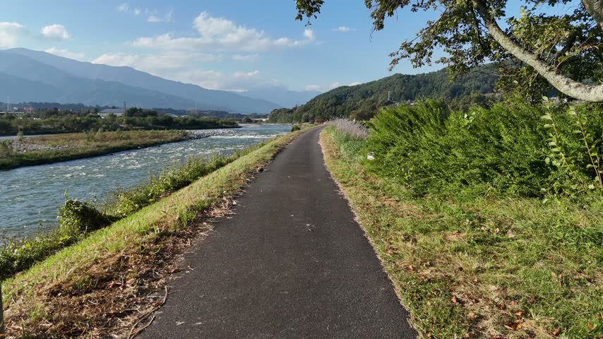 An asphalt trail follows a river beside tall grass and leafy shrubs toward mountain slopes beneath scattered clouds.