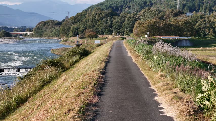 A narrow paved trail runs along grassy riverbanks beside a fast moving stream with trees and hills beyond.