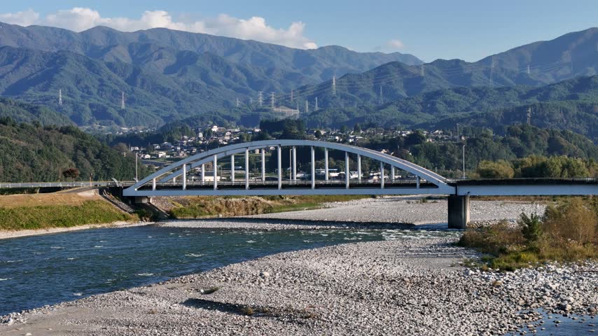 A white metal arch spans shallow water and gravel deposits with a town below a forest covered mountain ridge.