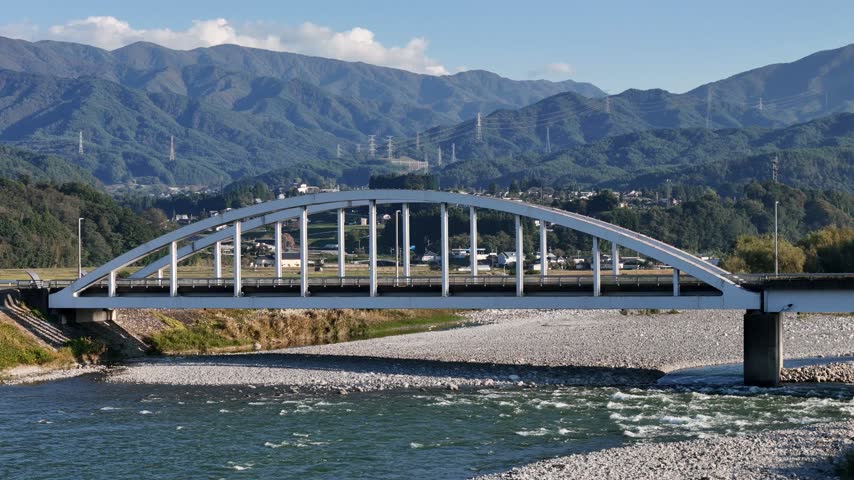 A white arched bridge crosses a pale gravel riverbed with green hills and a rural settlement behind.