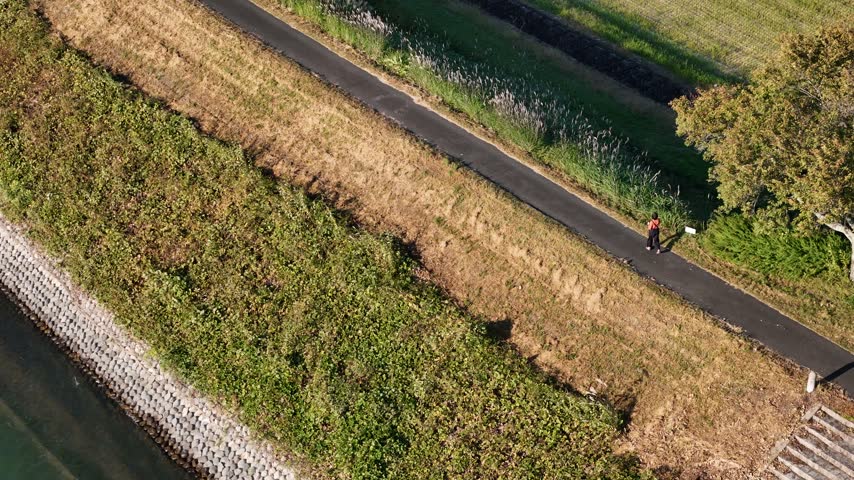 An overhead view shows one person on a paved riverside trail bordered by sloped grass, shrubs and a stone embankment.