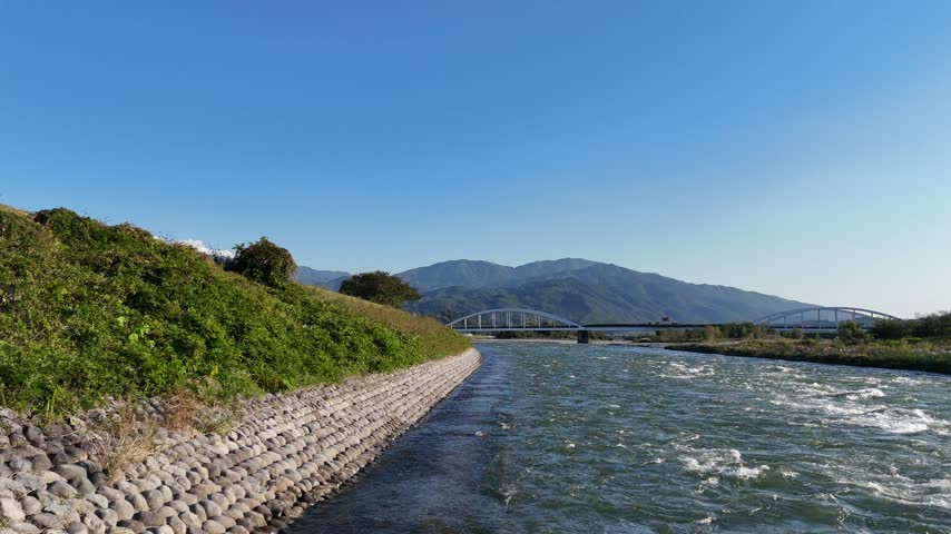 A low view follows flowing river water beside a concrete block embankment toward distant white arched road bridges.