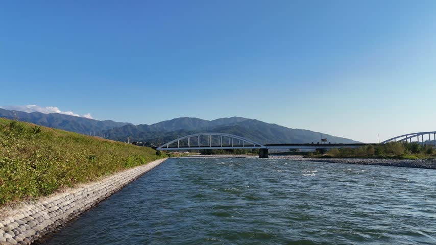 A low river view follows current past a stepped embankment toward a pale road bridge with a mountain ridge beyond.