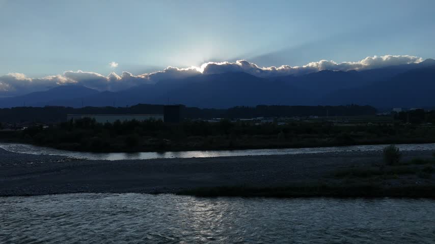 A broad river lies in shadow below jagged mountain silhouettes traced by sunlit clouds along the horizon.