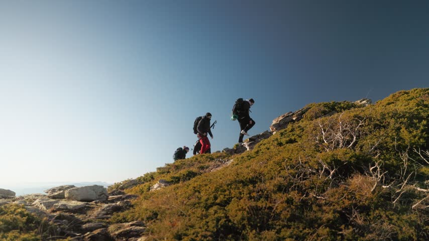 A tracking shot follows hikers climbing a sunny ridge with poles and backpacks.