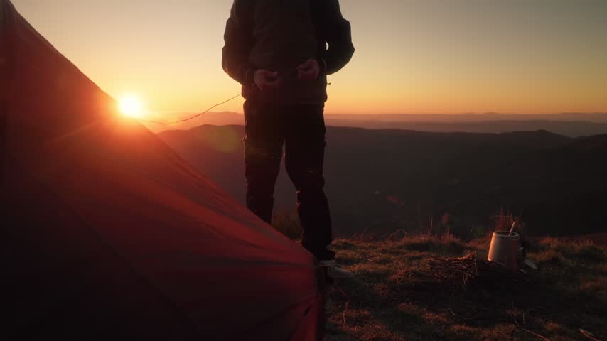 A silhouetted hiker secures tent stakes beside a mountain camp at sunset.