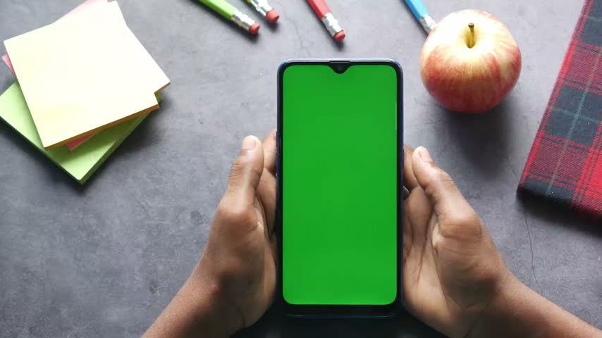 Top view shot of hands holding vertical smartphone with chroma key green screen background surrounded by office desk supplies like sticky notes pencils and apple for mockup presentation.