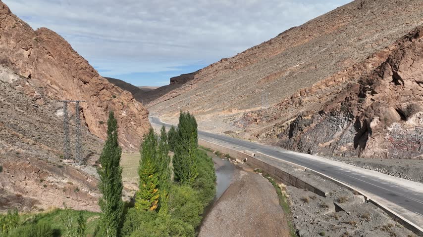 Aerial view of a paved road winding through a narrow rocky mountain valley beside a small watercourse and tall green trees in Morocco