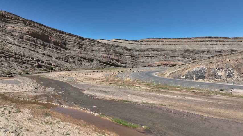 Wide view of horizontally layered rocky cliffs, a winding paved road and a shallow stream crossing a gravel valley in Morocco