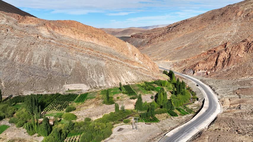 Aerial view of a winding paved road curving around eroded rocky mountains beside green agricultural fields in Morocco