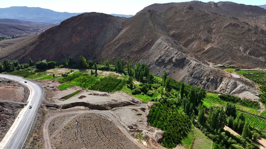 Aerial view of steep mountains with tilted rock layers overlooking green orchards, cultivated fields and a winding road in Morocco
