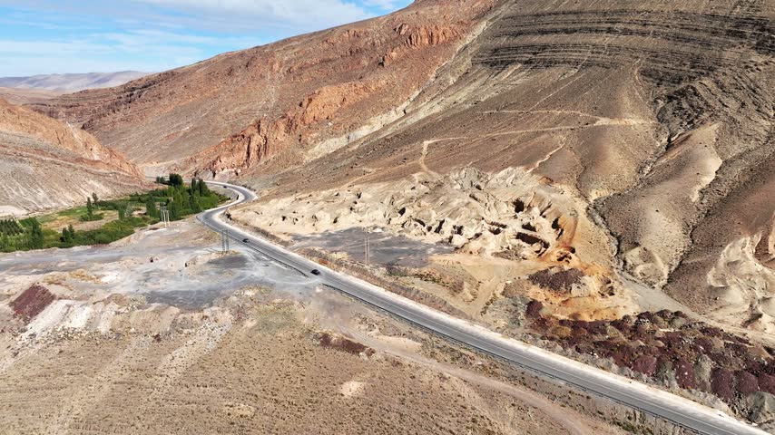 Aerial view of a winding paved road crossing a dry rocky valley beneath steep, layered mountain slopes in Morocco