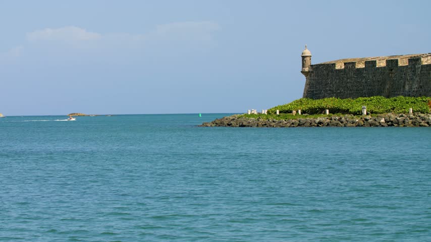 A view of one of the Garita lookouts at Fort San Felipe del Morro in San Juan, Puerto Rico under clear blue skies. Stoned fortified wall construction as small vessel travels along the bay.