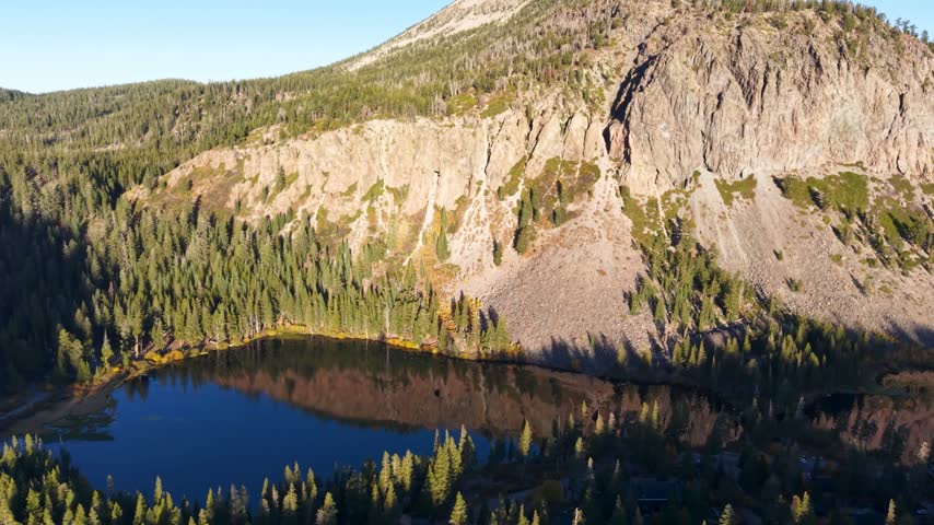 Aerial pull away shot of Twin Lakes in Mammoth Mountain California. Dramatic Flight away from the mountain face reveals a glassy blue lake in the early Autumn morning.