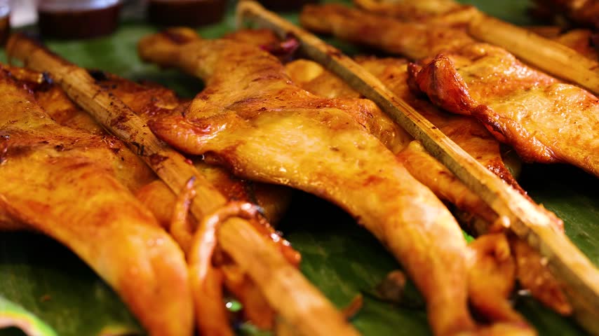 Close up shot of roasted chicken meat on bamboo sticks served on fresh banana leaf in traditional outdoor Asian market stall.
