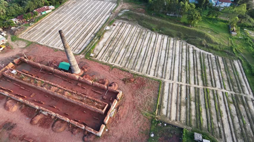 Drone orbits around a disused traditional brick kiln with a tall chimney and empty open chambers, surrounded by drying brick fields, red earth and green trees, Bangladesh, 4K.