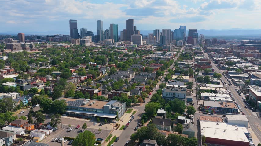 Denver, Colorado, panoramic summer cityscape from above shows leafy neighborhoods, parks, rows of houses, and modern skyscrapers, framed by the distant Front Range under a partly cloudy sky.