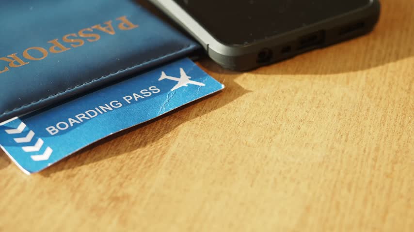 Travel preparation concept showing passport cover with boarding pass, smartphone, pen and notebook placed on wooden desk, closeup slow panning shot of flight essentials.