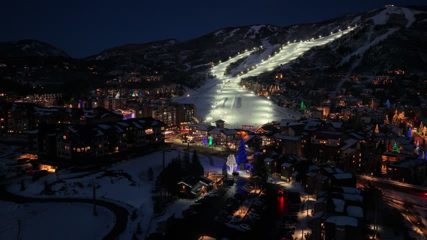 Winter night view resembling Deer Valley, Utah shows bright ski runs descending toward a snow covered town, with active night skiing, illuminated lodges, roads, and neighborhood lights.
