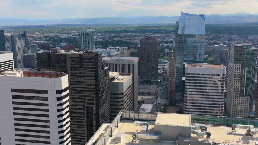 Close aerial drone view of downtown Denver, Colorado, with office towers, the historic Daniels and Fisher clock tower, city streets, and the Rocky Mountains stretching across the horizon.