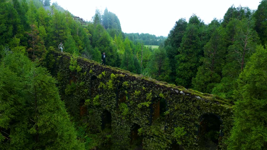 Two people walk across a mossy stone aqueduct surrounded by lush forest in spring