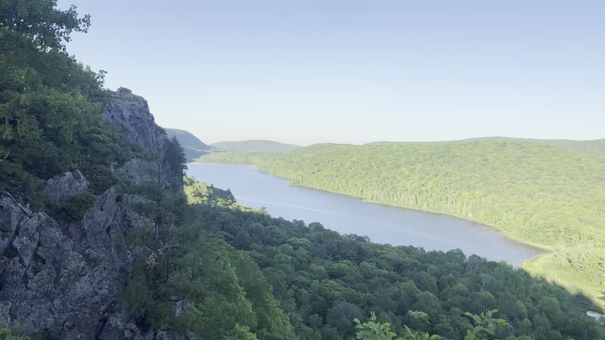 A rightward pan from a rocky overlook reveals Lake of the Clouds surrounded by green forest and ridges in Porcupine Mountains Wilderness State Park near Ontonagon Michigan.