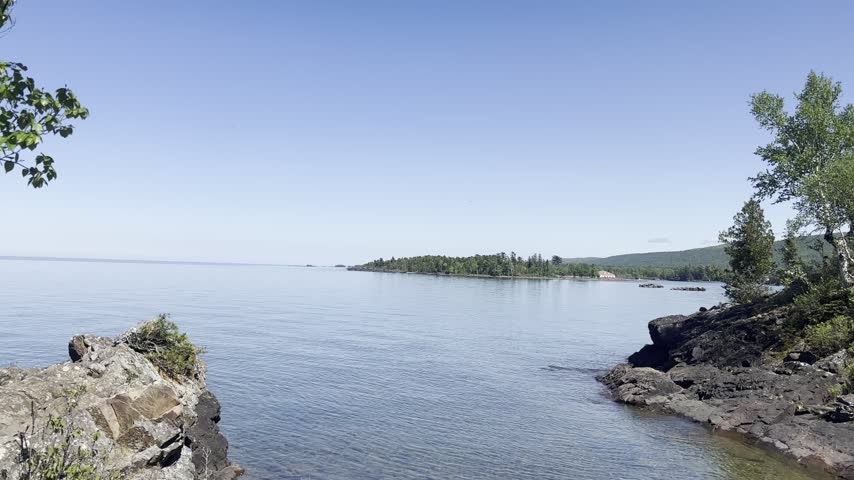 A coastal pan reveals Eagle Harbor Lighthouse above rugged shoreline rocks with calm Lake Superior water leafy trees and a forested headland in Michigan.