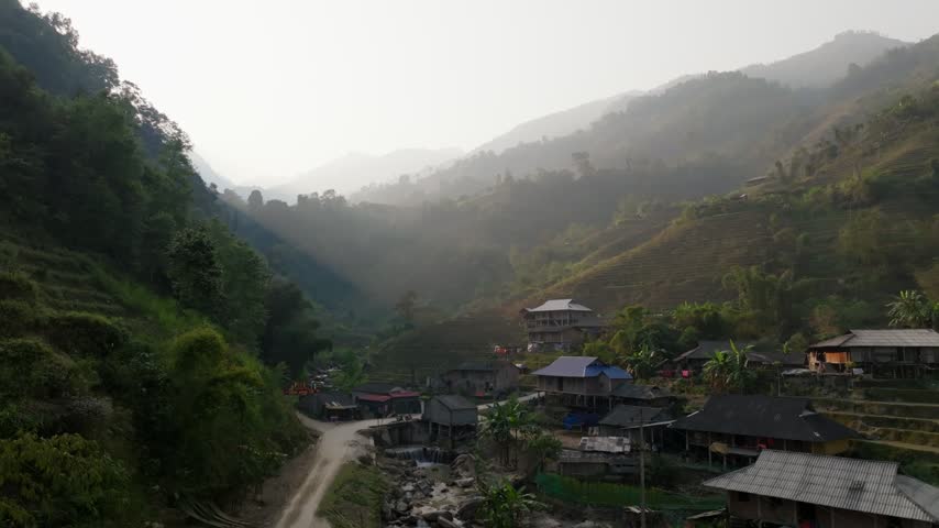 Drone rises past dark green trees to reveal a village with blue and grey roofs and rice terraces in a misty valley in Hoàng Su Phì, Vietnam, afternoon backlight.