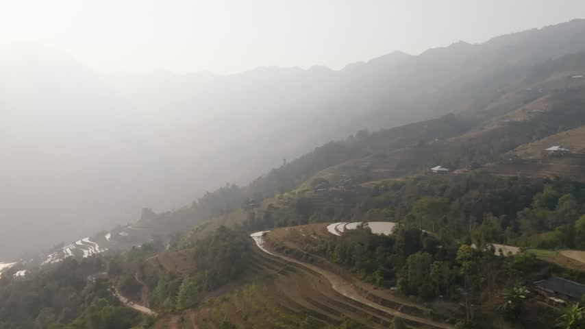 Aerial view of a winding road and water-filled terraces on a forested hillside in thick backlit haze over a valley in Hoàng Su Phì, Vietnam, late afternoon.