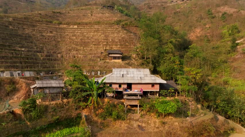Aerial view pulling back from a lone pink house beside a forested gully, with large fallow terraces on the slope behind in Hoàng Su Phì, Vietnam, late sun.