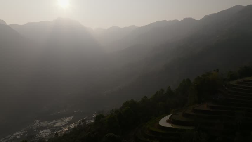 Aerial view rising over a dark ridge to a vast hazy valley with flooded terraces glinting below sun rays over distant peaks, Hoàng Su Phì, Vietnam, sunset.