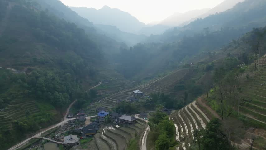 Aerial view descending toward blue-roofed houses and layered rice terraces on a forested slope in Hoàng Su Phì, Vietnam, with a misty valley behind in soft haze.