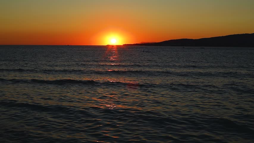 Sunset over calm sea waters with boats visible on the horizon, reflecting warm orange and yellow hues against the tranquil sea surface at dusk
