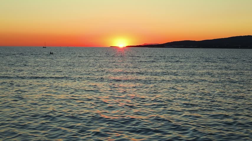Sunset over calm sea waters with boats visible on the horizon, reflecting warm orange and yellow hues against the tranquil sea surface at dusk