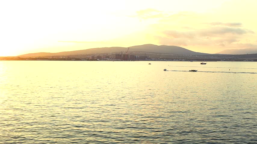 Sunset over calm water with boats gliding across the surface, silhouetted against the vibrant orange and yellow sky, creating a serene coastal scene