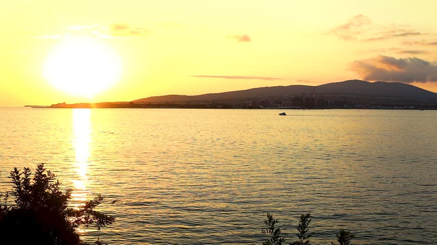 Sunset over calm water with boats gliding across the surface, silhouetted against the vibrant orange and yellow sky, creating a serene coastal scene