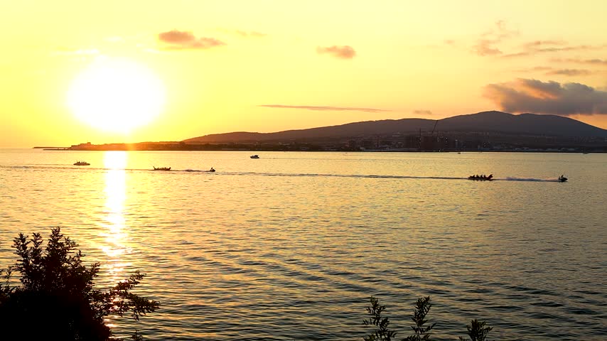 Sunset over calm water with boats gliding across the surface, silhouetted against the vibrant orange and yellow sky, creating a serene coastal scene