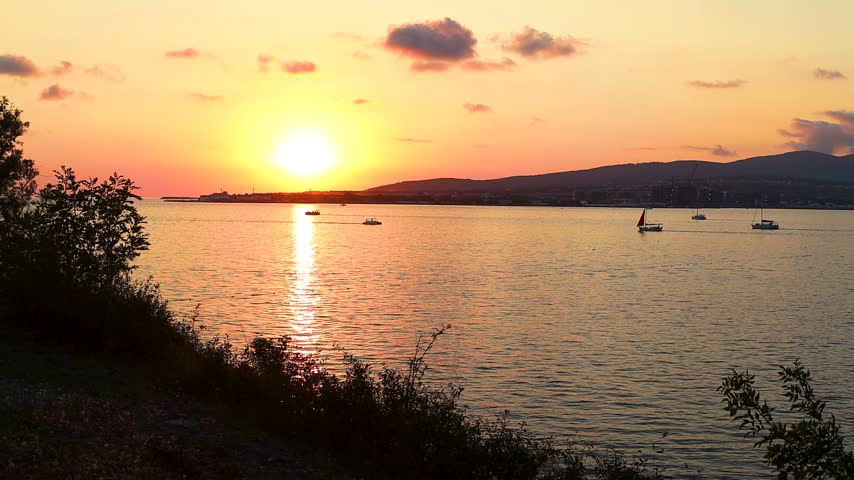 Sunset over calm water with boats sailing, silhouetted trees framing the scene, and distant hills creating a serene landscape in a coastal region