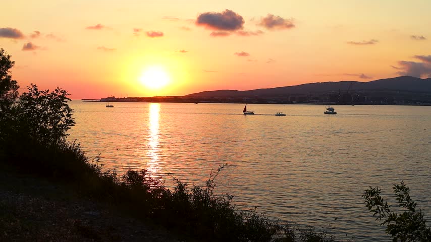 Sunset over calm water with boats sailing, silhouetted trees framing the scene, and distant hills creating a serene landscape in a coastal region