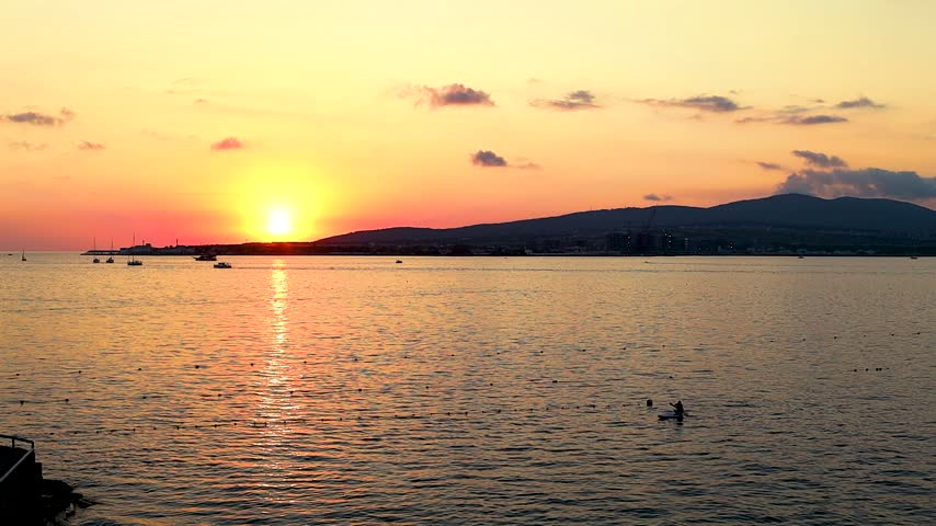 Sunset over calm water with boats sailing, silhouetted trees framing the scene, and distant hills creating a serene landscape in a coastal region