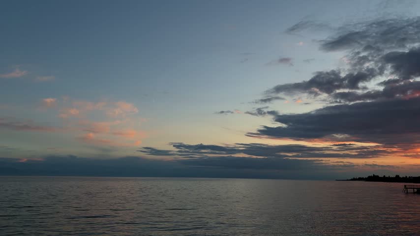 Slow pan across calm lake at dusk with crescent moon, dark clouds and orange afterglow over pier silhouette, Issyk-Kul, Kyrgyzstan