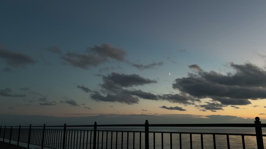 Crescent moon among dark clouds in twilight sky over calm lake with silhouetted pier railing in foreground, Issyk-Kul, Kyrgyzstan