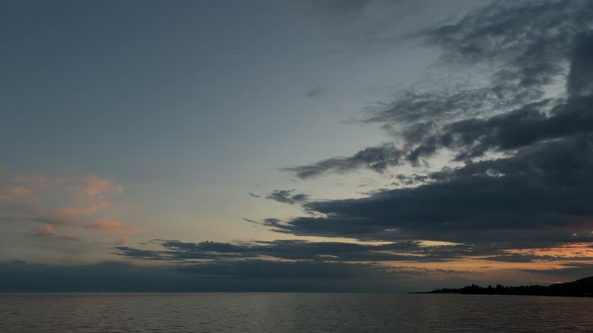Helicopter silhouette flying past thin crescent moon over calm mountain lake at dusk with dark clouds and afterglow, Issyk-Kul, Kyrgyzstan