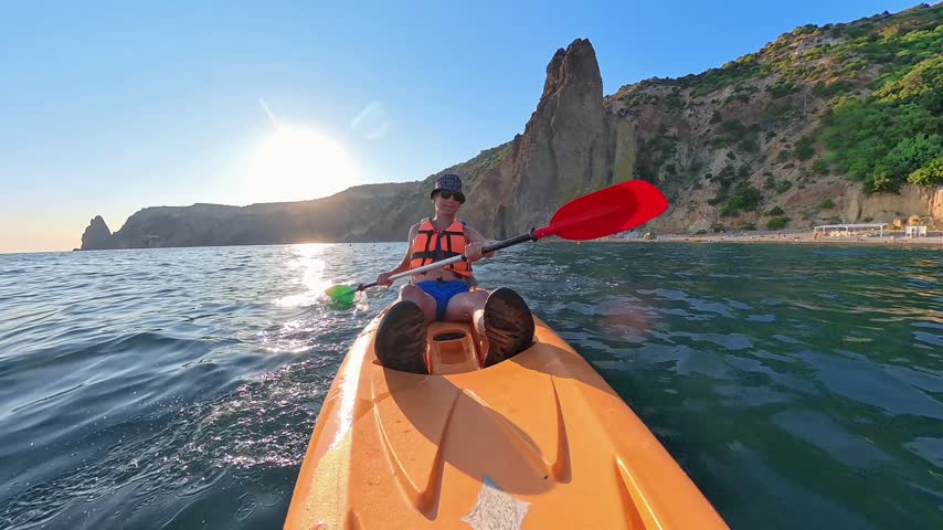 An active man adventurer paddles a bright orange kayak through coastal waters alongside steep rocky precipices. Brilliant sunlight shimmers on the sea surface as she navigates forward in a point-of-view follow capture