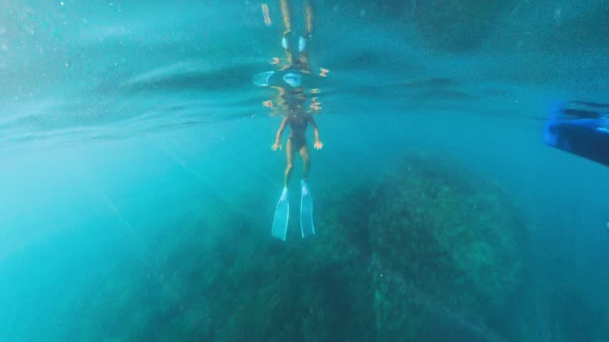Woman wearing long fins freediving in clear turquoise sea water near rocky reefs. Split-level over-under perspective capturing her surface float and underwater descent