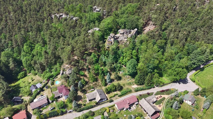 Drone captures tranquil rural scene showing clustered homes and cottages along curved road, surrounded by lush forest, gardens, meadows, and open farmland under bright blue sky during late spring.